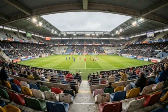 Vue grand angle des tribunes d'un stade de Ligue 1 à budget modeste lors d'un jour de match, avec pelouse impeccable et affluence partielle dans les gradins